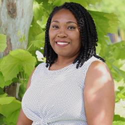 Photo of Latoya Lewis outside in front of a tree. She is wearing a black and white printed tank top style blouse. Latoya has short curly dark hair. 