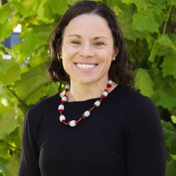 Photo of Laura Fogle outside in front of a tree. She is wearing a black long sleeve shirt with a red and white beaded necklace. Laura has short curly dark brown hair. 