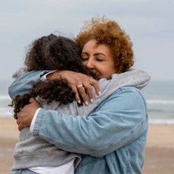 two women hugging on a beach. One is wearing a jean jacket the other a grey sweatshirt. It looks like a cold and cloudy day.