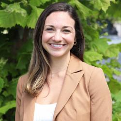 Photo of Danielle Trauth-Jurman outside in front of a tree. She is wearing a white blouse with a beidge blazer. Danielle has long dark brown hair blone highlights