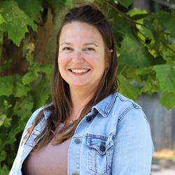 Photo of Naomi Zeiset outside in front of a tree. She is wearing a beige top under a light colored denim jacket. Naomi has long straight dark brown hair.