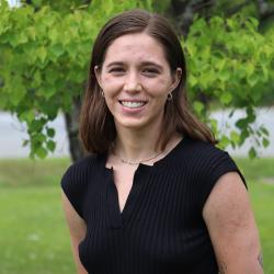 Photo of Natalie, standing outside in front of a tree. Natalie has shoulder length brown hair and is wearing a black, ribbed, sleeveless top