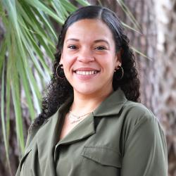 Photo of Allison Hilton, smiling, outside. She has long dark curly hair and wearing a green buttoned up top.