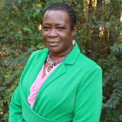 Photo of Regina Armistad. She has short hair, wearing a pink top, green blazer and gold earrings and a beaded necklace with large gold charm