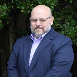 Andrew is wearing glasses, a light colored button up top and a navy blue jacket. He is smiling looking at the camera standing outside.