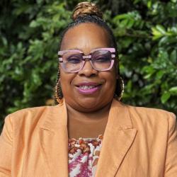 Denise Bryant is wearing glasses, gold hoop earrings, a floral top and peach colored jacket. She is looking at the camera smiling.
