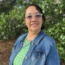 Karen Chew is standing outside, wearing glasses, a green patterned dress and denim jacket. She has dark hair, and is smiling at the camera