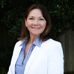 Piper has short brown hair, wears a white blazer and blue striped button up top. She is smiling at the camera.