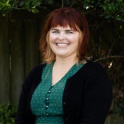 Photo of Nicole Krokosz. She has shoulder length red blonde hair and is wearing a green top and black jacket, smiling at the camera