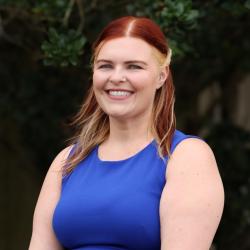 Photo of Nicole Krokosz. She has shoulder length red blonde hair and is wearing a bright blue dress, smiling at the camera