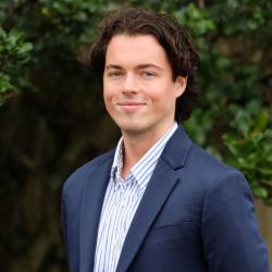 Photo of Will Williams. He has short dark hair, wearing a blue and white shirt and blue jacket, smiling at the camera