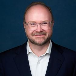 A photo of DJ standing in front of a dark blue background. DJ is smiling at the camera and wearing glasses, a navy suit coat, white collared shirt and no tie.