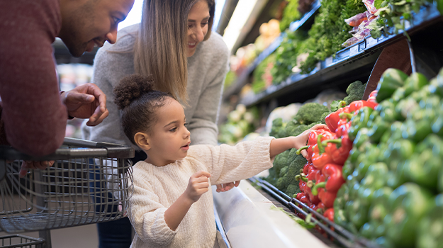 A family of three at a grocery store. A child looks at the produce section and is selecting a fresh vegetable