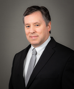 Chris Glenn is wearing a black blazer, grey tie, and white shirt. He is smiling in front of a grey background for his employee photo.