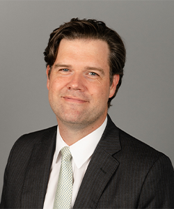 Peter is wearing a black blazer, white shirt, and green tie. He is smiling in front of a grey background for his employee headshot.