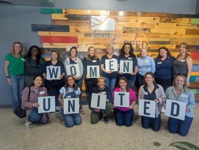 Group photo of Women United members at the Lowcountry Food Bank. They are posed for the photo, each hold a letter that spells out Women United.