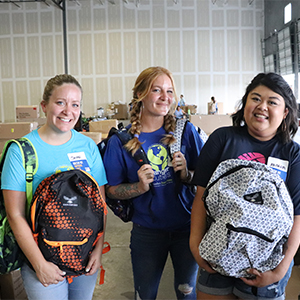 Three women pose for a photo holding backpacks with several boxes behind them in a warehouse setting