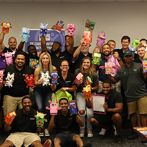 A large group of volunteers pose for a fun and silly photo holding up paper hand puppets that were made during a volunteer kit assembly event.