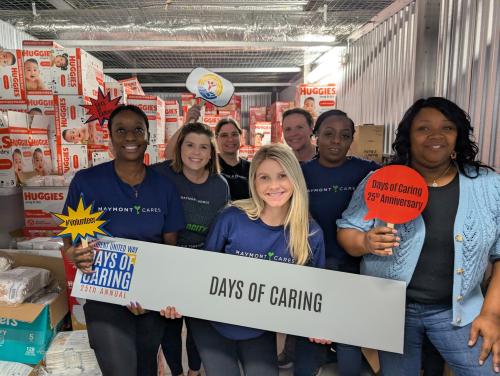 A group of individuals stand in front of a storage space filled with diapers. They are holding a sign that reads Days of Caring.