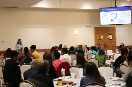 A full room with people sitting around tables, a person stands at the head of the room speaking into a podium mic