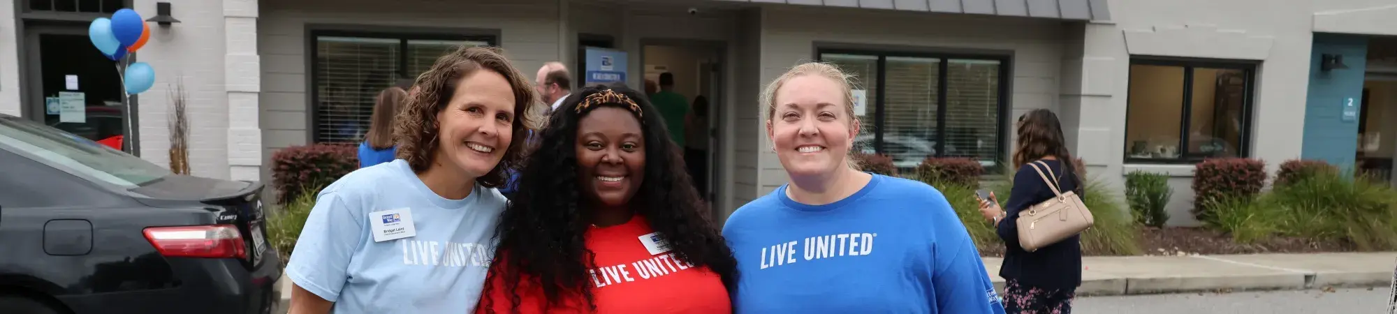 Three women stand in front of the Dorchester Resource Connection Center. They are smiling at the camera and each wearing a different color shirt that says LIVE UNITED