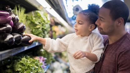 A man and child are standing next to the produce section of a grocery store selecting an eggplant.