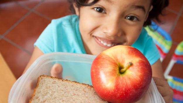 Young girl holding a tupperware container with a sandwich and apple in it. The girl is smiling, and appears to be in a school.