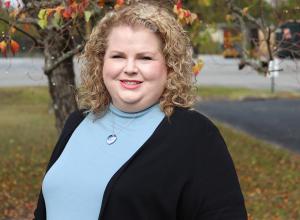 Headshot photo of Andrea Boccucci in front of a tree. She is wearing a light blue turtle neck with a black cardigan. Andrea has short curly blonde hair. 