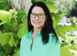 Photo of Louise Lawton outside in front of a tree. She is wearing a mint green button down shirt, black glasses and gold hoop earrings. Louise has shoulder length straight dark hair.